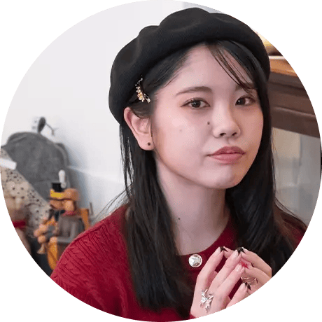 黒いベレー帽と赤い服を身に着けた女性が、室内で手を組んでカメラを見つめている