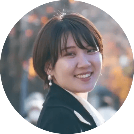 短髪で黒いコートを着た女性が、屋外で振り向きながら微笑んでいる秋の風景