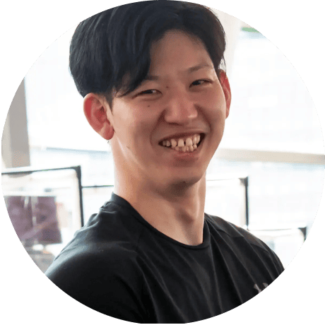 黒いTシャツを着た男性が、室内で笑顔を見せながらカメラに向かっている