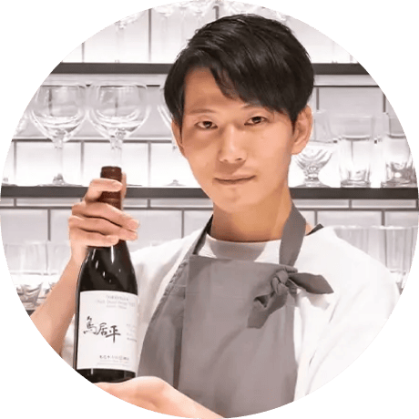 グレーのエプロンを着た男性が、ガラス棚の前で日本酒の瓶を手に持ちポーズを取っている